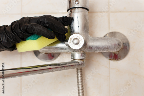 Limescale crust on bathroom mixer faucet. Hands in disposable rubber (latex or vinyl) gloves cleaning hard water calcium carbonate deposit on sink or bath tap. Dirty plumbing covered with lime scale