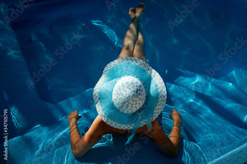 beautiful young woman in wide-brimmed blue hat sunbathing lying in the pool with turquoise clear water