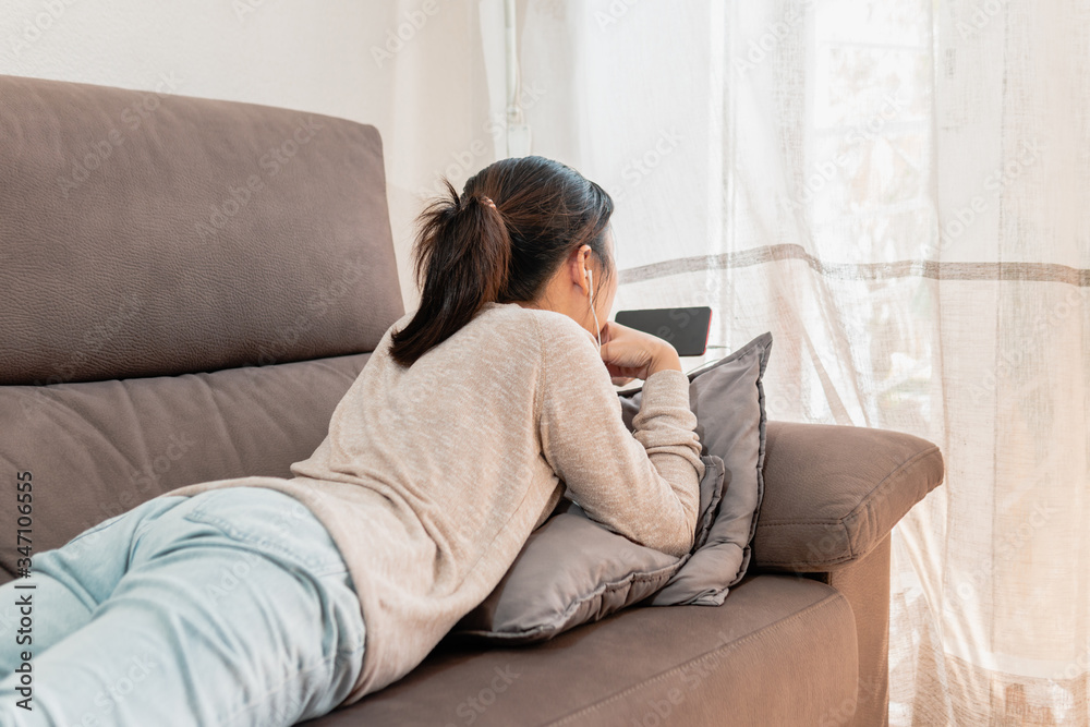 Asian woman relaxing at home lying on the couch watching videos on a phone wearing earphones. Girl taking a break resting on the sofa of a cozy house. Asian woman relaxing at home lying on the couch watching videos on a phone wearing earphones. Girl taking a break resting on the sofa of a cozy house.