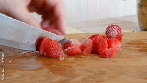 Female hands cut a frozen strawberry with a knife for future fruit salad or smoothie on a wooden kitchen board. Morning breakfast at home. photo