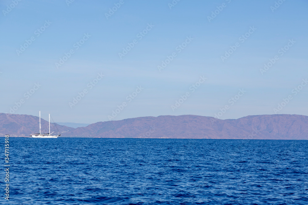 A single white sailing boat is traveling in the blue Aegean sea with mountain and sky background and with copy space.