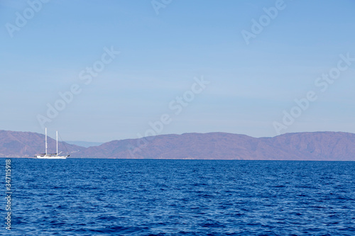 A single white sailing boat is traveling in the blue Aegean sea with mountain and sky background and with copy space.