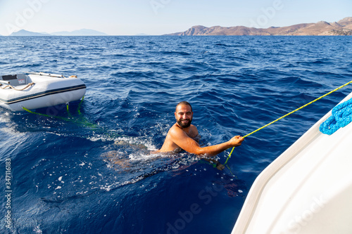 Young smiling mediterranean man with black beard, holding a rope behind the sailing boat and endjoying swimming in blue waters. Inflatable boat, sky and rocky islands in background © Stockwars