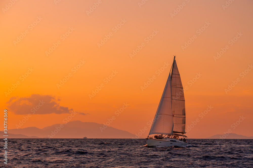 Sailing boat floating on the blue Aegean Sea during sunrise / sunset with open main sail and genoa. Orange sky with clouds and islands in background for copy space