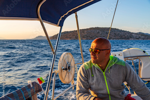 Bald man with sun glasses is sitting on deck of sailing boat and enjoying the summer and his journey. View from stern to blue waters, sky and green island. © Stockwars