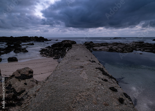 Low angle shot straight along concrete jetty to ocean at dusk with rock pools and reflections on water. Long exposure.