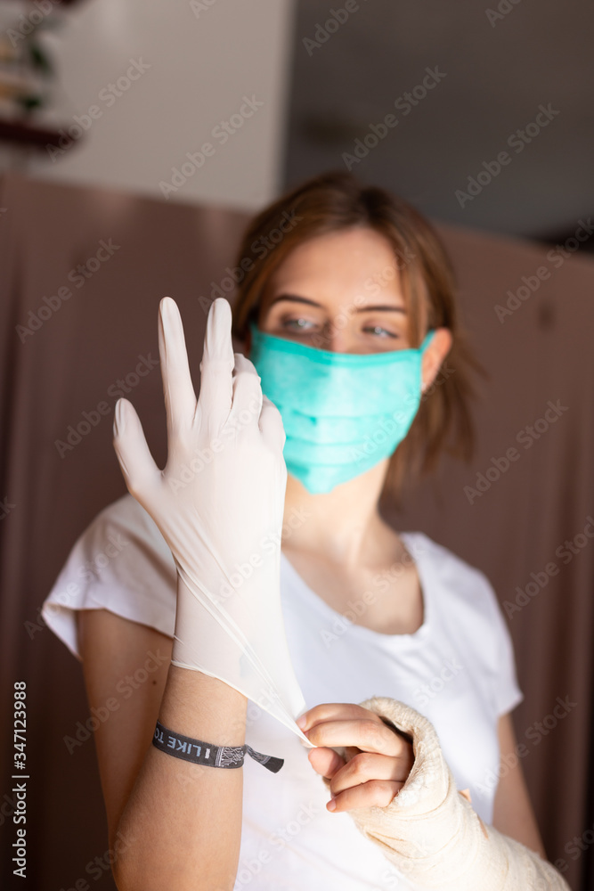 portrait of beautiful young woman wearing cotton green mask and white medical/surgical gloves on pastel background. She is adjusting gloves.