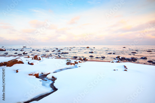 A view of the snow-covered Baltic sea coast at sunset. Stones in the water close-up. Clear blue sky with colorful clouds. Stunning cloudscape. Warm evening light. Idyllic winter scene. Kaltene, Latvia photo