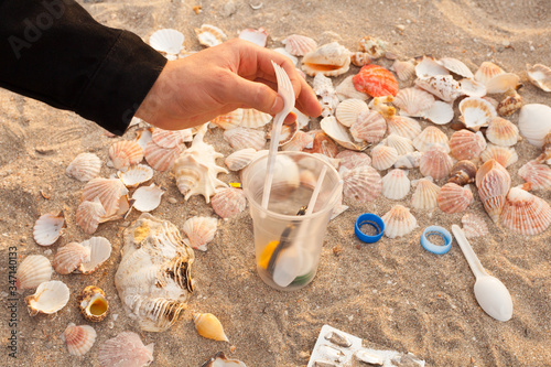 A man's hand collects and removes plastic waste on the beach: bags, disposable plastic appliances, plastic lids among the shells. 