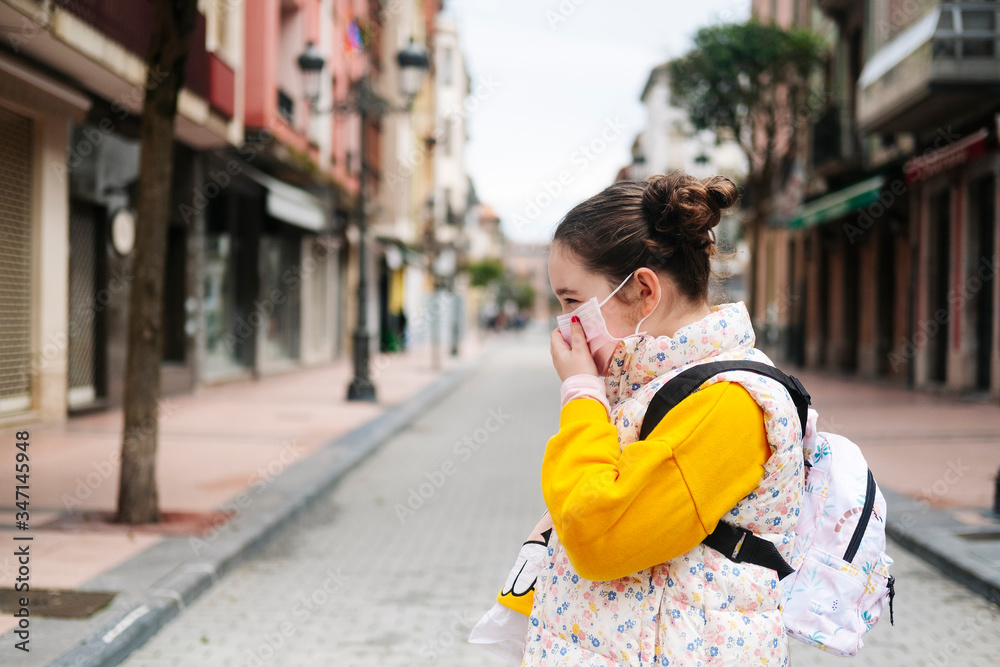 A girl with a mask on her face in the street of a city. He wears a yellow sweater and backpack