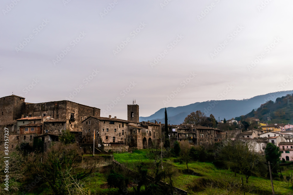 A panoramic view of Santa Pau (Girona, Spain), its old town, fortress, and other building, with the Pyrenees mountains in the background, in the cloudy afternoon in winter