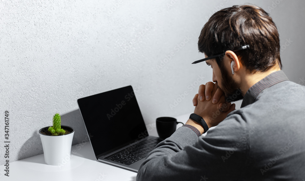 Back view of young thoughtful businessman with pencil on ear, looking in laptop, using wireless earphones, wearing smartwatch, coffee cup and cactus on white desk. Background of grey textured wall. Back view of young thoughtful businessman with pencil on ear, looking in laptop, using wireless earphones, wearing smartwatch, coffee cup and cactus on white desk. Background of grey textured wall.