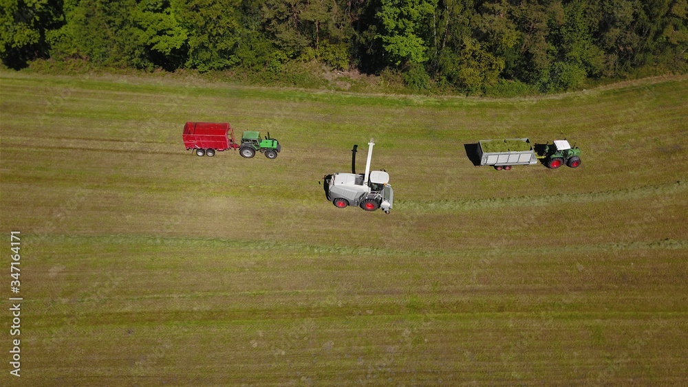 Obraz premium Landwirtschaft: Feldhäcksler, Traktor mit Ladewagen bei Aufnahme von Gras für Grassilage auf einer gemähten Wiese, Vogelperspektive