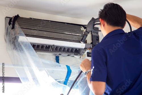 The technicians are cleaning the air conditioner by spraying water. Hand and water spray are cleaning the air conditioner on with white wall background.