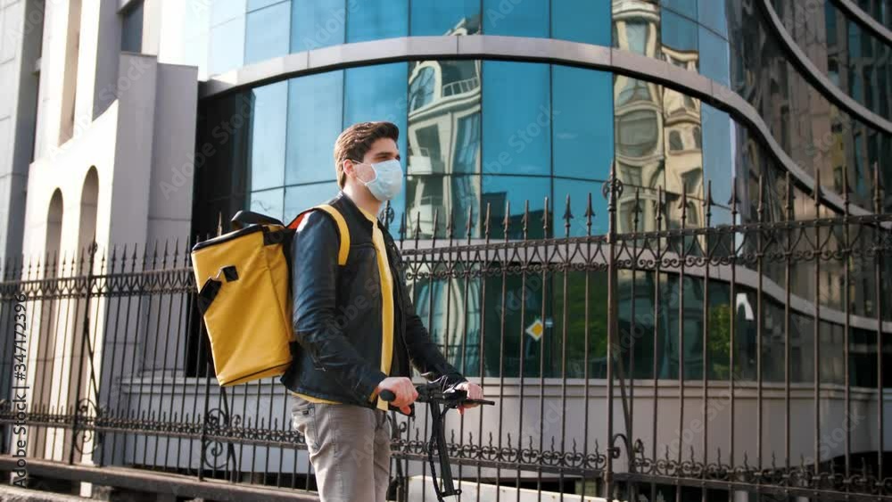 Delivery man with yellow bagpack and protective mask riding electric scooter through the city with food delivery, business center background Delivery man with yellow bagpack and protective mask riding electric scooter through the city with food delivery, business center background