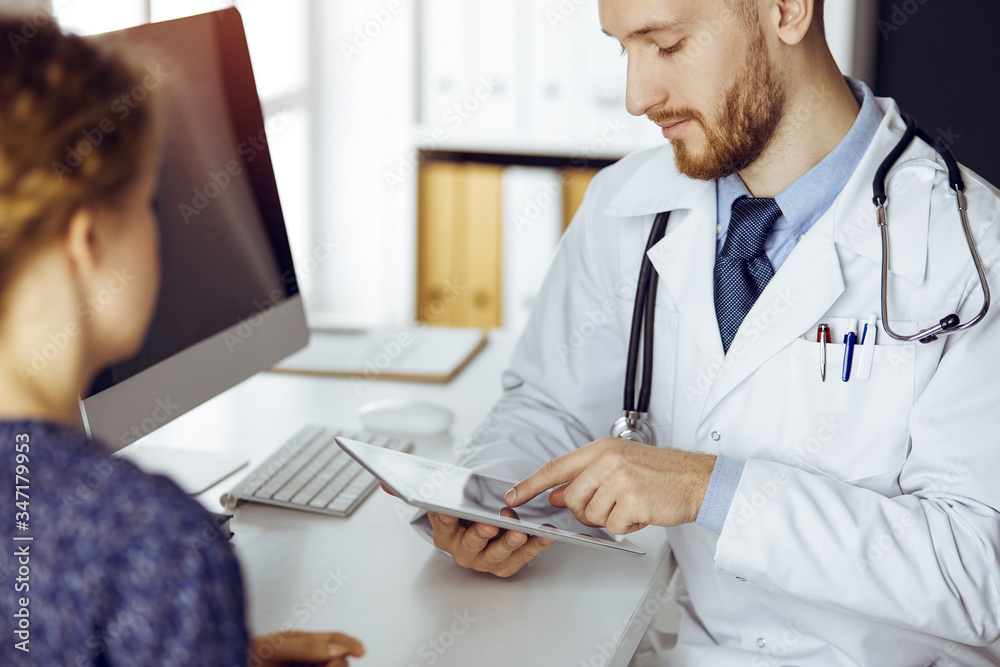Friendly red-bearded doctor and patient woman discussing current health examination while sitting and using tablet computer in clinic. Medicine concept Friendly red-bearded doctor and patient woman discussing current health examination while sitting and using tablet computer in clinic. Medicine concept
