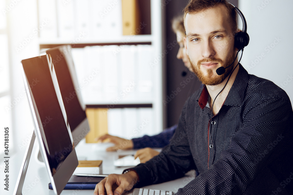 Fototapeta premium Red-bearded businessman talking by headset near his female colleague while sitting in modern sunny office. Diverse people group in call center
