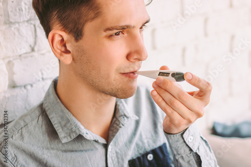 Closeup ill hipster man student putting digital thermometer in mouth for body temperature check. Sick businessman freelancer use electronic thermometer. Measuring temperature at home. Flu prevention