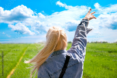 A young girl is looking at the field. Beautiful blue sky. Background green field and sky. The blonde reaches for the sky with her hand in a denim jacket. Beautiful girl admires nature.