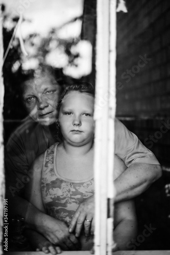 Black and white dramatic mood portrait of grandmother hugs her granddaughter. Different generations. Old and young. Strange people looking through window. Rustic family indoor. Parents care children.