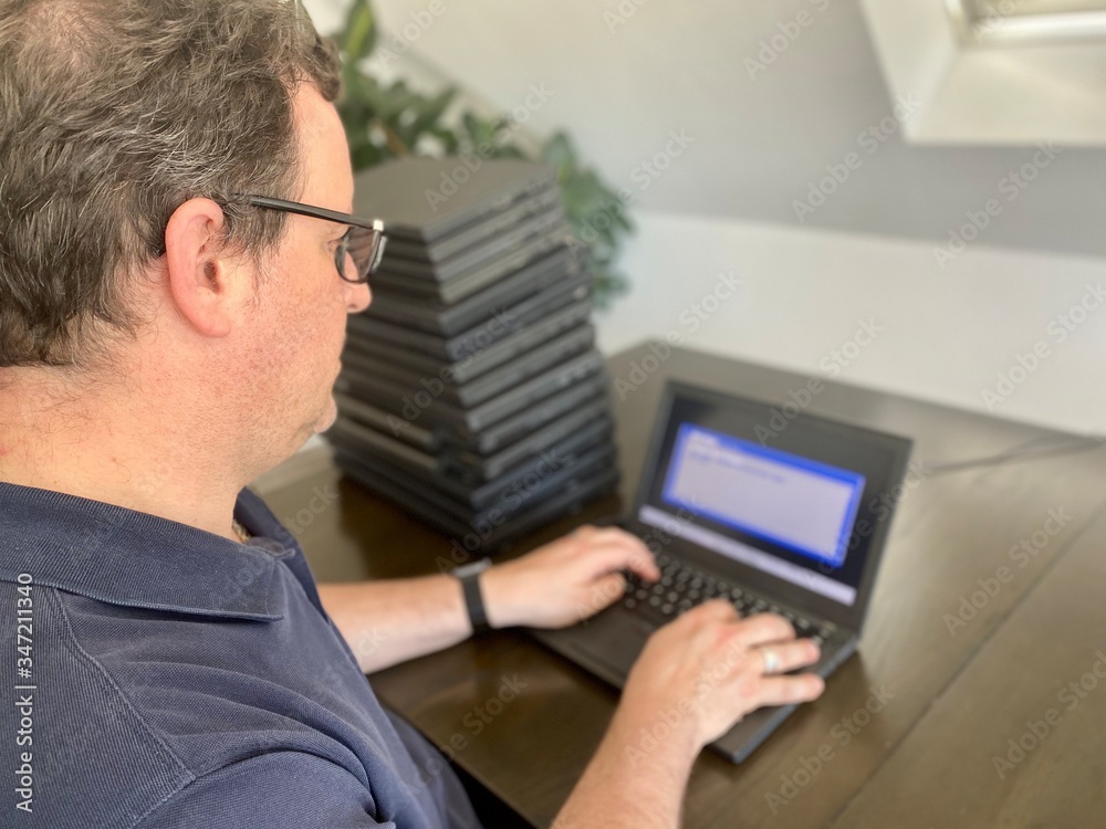 Corona Crisis: A casually dressed caucasian middle aged man is installing a laptop computer with a stack of several more computers next to him. Laptops are used and second hand, might be for people in Corona Crisis: A casually dressed caucasian middle aged man is installing a laptop computer with a stack of several more computers next to him. Laptops are used and second hand, might be for people in