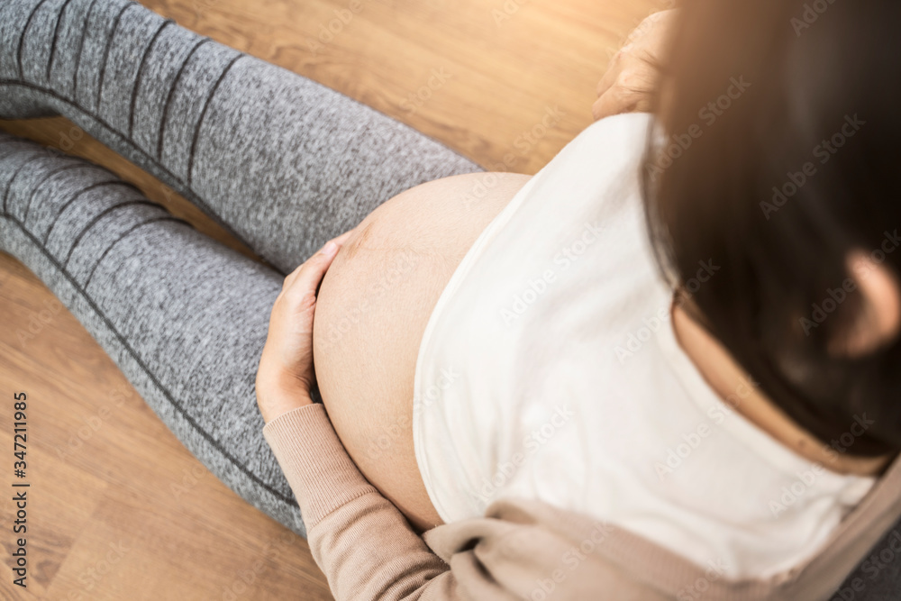beautiful asian pregnant woman placing hands on baby lump feeling heartbeat smiling joyfully, sitting against sofa relaxing resting from tiredness, in living room with brick texture