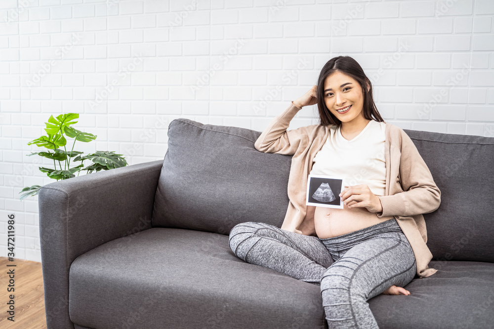beautiful asian pregnant woman sitting on sofa holding ultrasound baby picture and smiling with happiness, resting relaxing in living room from hormone stress, wearing comfy stretch pants and jumper beautiful asian pregnant woman sitting on sofa holding ultrasound baby picture and smiling with happiness, resting relaxing in living room from hormone stress, wearing comfy stretch pants and jumper