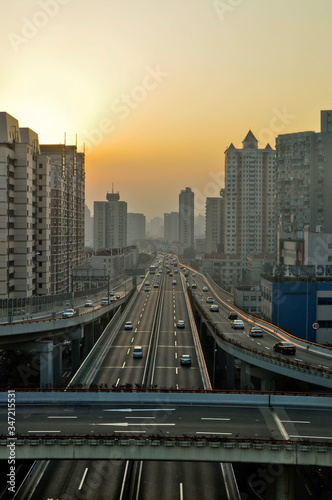 Evening mood in Shanghai between skyscrapers. The sun sets in the background in the smog. The elevated roads bridges lead the Traffic that never stops. The sky is an orange yellow with haze.
