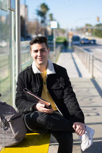 Young student sitting cross-legged at the bus stop holding a notebook while looking at the camera with a smile