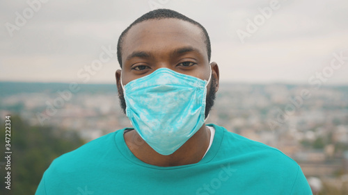 Pandemic 2020. Portrait of african healthy man wearing protective face mask and goggles looking straight standing on the hilltop above city. Quarantine. Safety care.