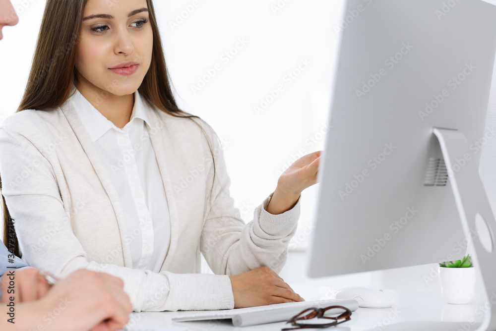 Young woman sitting at the desk with computer in white colored office. Looks like student girl or business lady communicating with casual dressed man Young woman sitting at the desk with computer in white colored office. Looks like student girl or business lady communicating with casual dressed man