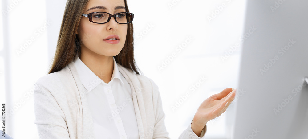 Young woman sitting at the desk with computer in white colored office. Looks like student girl or business lady communicating with casual dressed man Young woman sitting at the desk with computer in white colored office. Looks like student girl or business lady communicating with casual dressed man