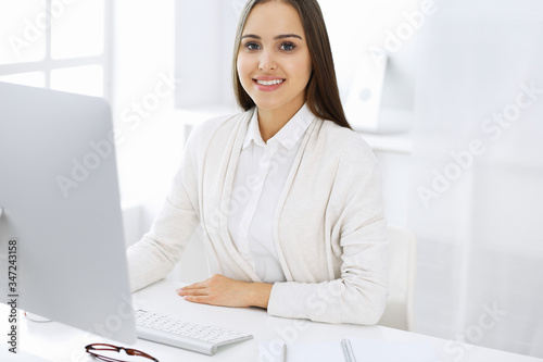 Young cheerful woman sitting at the desk with computer and looking at camera in white colored office. Looks like student girl or business lady. Headshot