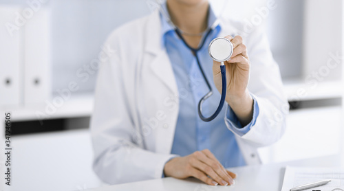 Unknown woman-doctor in blue blouse is holding stethoscope head while sitting at the desk in clinic, close-up. Physician ready to examine and help patient. Medicine concept