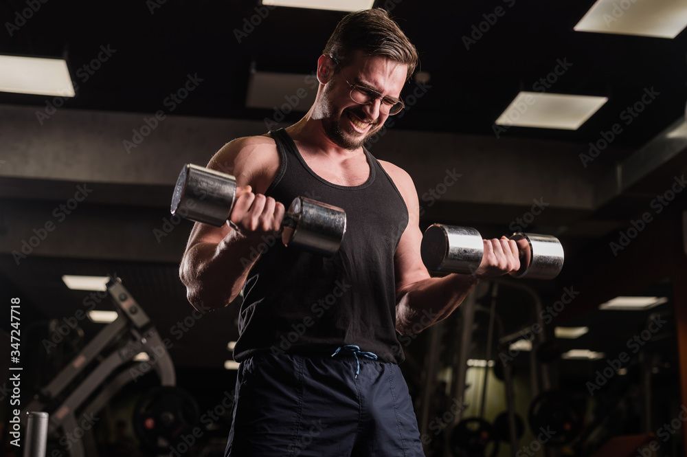 A man with an effort does an exercise for biceps and triceps with a dumbbell. Embossed bodybuilder smiles. Muscular guy with glasses in the gym. A man with an effort does an exercise for biceps and triceps with a dumbbell. Embossed bodybuilder smiles. Muscular guy with glasses in the gym.
