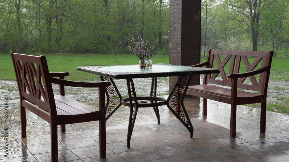 Spring heavy rain pours down on the street splattering the tiles on the veranda on which there are two benches and a table decorated with a bouquet Spring heavy rain pours down on the street splattering the tiles on the veranda on which there are two benches and a table decorated with a bouquet