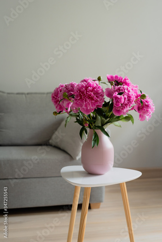 Blooming Pink peony flower with green leaves in pink vase on the white table, in the room, grey background, lifestyle image, copy space