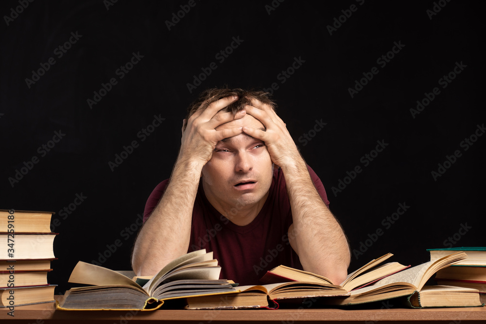 A young man was sitting at a table above books, holding his head in his hands. Piles of books are lying nearby. Black background. isolate. difficulties in education and quarantine. A young man was sitting at a table above books, holding his head in his hands. Piles of books are lying nearby. Black background. isolate. difficulties in education and quarantine.