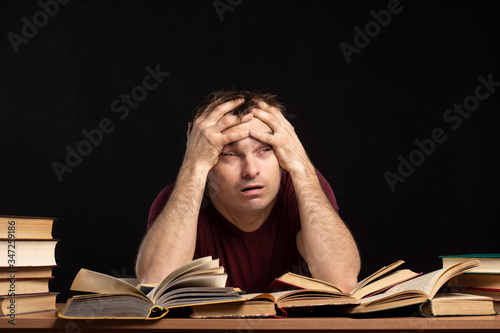 A young man was sitting at a table above books, holding his head in his hands. Piles of books are lying nearby. Black background. isolate. difficulties in education and quarantine.