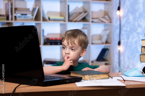 little boy sits at a table in the apartment and looks at the laptop. nibbling fingers on the background of a bookcase. difficulty learning online. distance learning online. © Олег Кононов