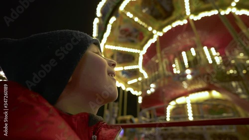 A boy looks at the illuminated carousel in the evening. Child near the whirling merry-go-round at an amusement park. Children Christmas carousel in garlands. Fun family vacation. Happy childhood.