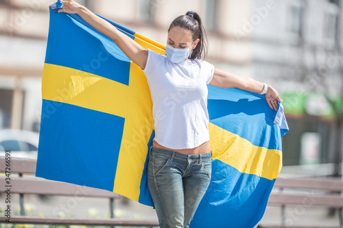 Brunetter standing outdoors looks at the Swedish flag she holds  with a face mask on