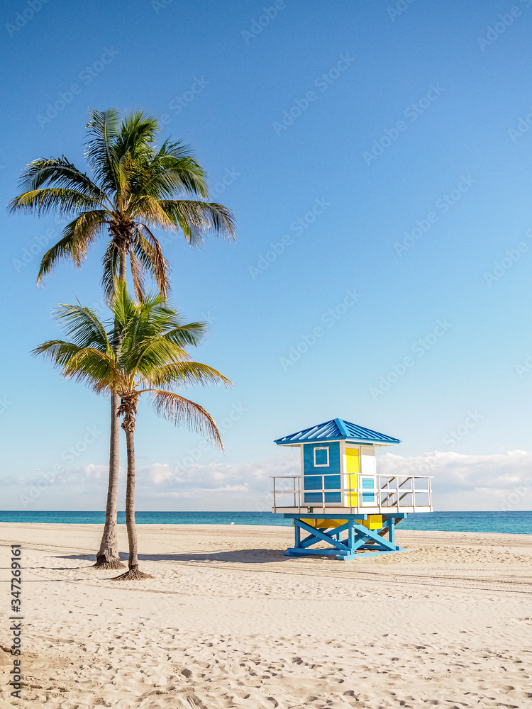 Naklejka premium Colorful blue and yellow lifeguard station on beach with palm trees and blue sky copy space.