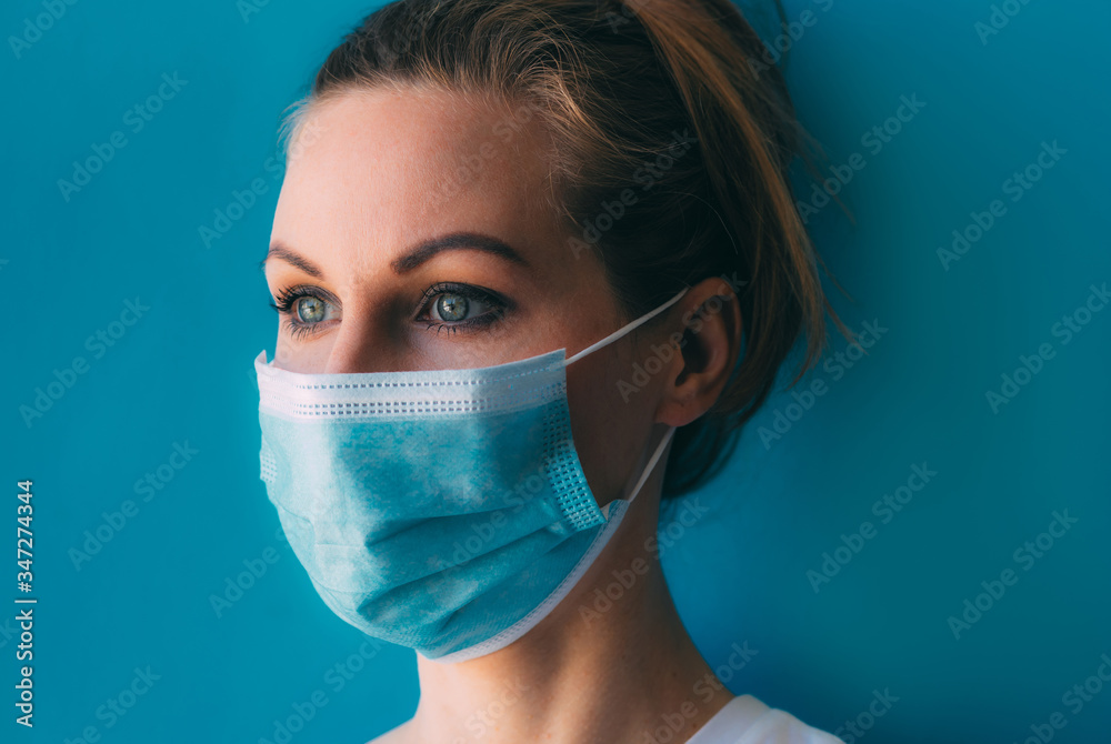 Portrait of young female doctor in medical mask and white gown on blue background. Female doctor in clinic in surgical mask with space for text. Head and shoulders portrait of intern in hospital Portrait of young female doctor in medical mask and white gown on blue background. Female doctor in clinic in surgical mask with space for text. Head and shoulders portrait of intern in hospital