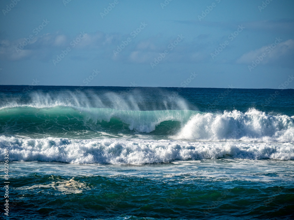 Big waves crash close to shore with aquamarine seas, white foam, sand and blue skies.