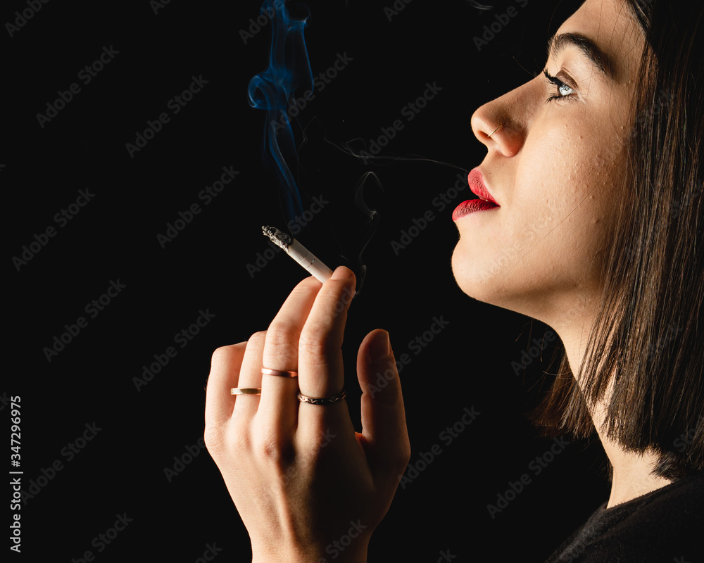 Obraz premium Low key close-up young woman smoking cigarette on black background. Copy space for text message.