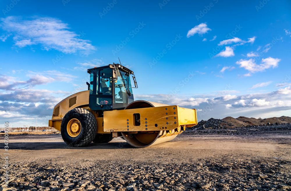 Eye catching yellow road roller with enclosed climate controlled cabin stands on not ready new road, stones, blue sky, clouds, left side view. Clean shiny old heavy tractor Eye catching yellow road roller with enclosed climate controlled cabin stands on not ready new road, stones, blue sky, clouds, left side view. Clean shiny old heavy tractor