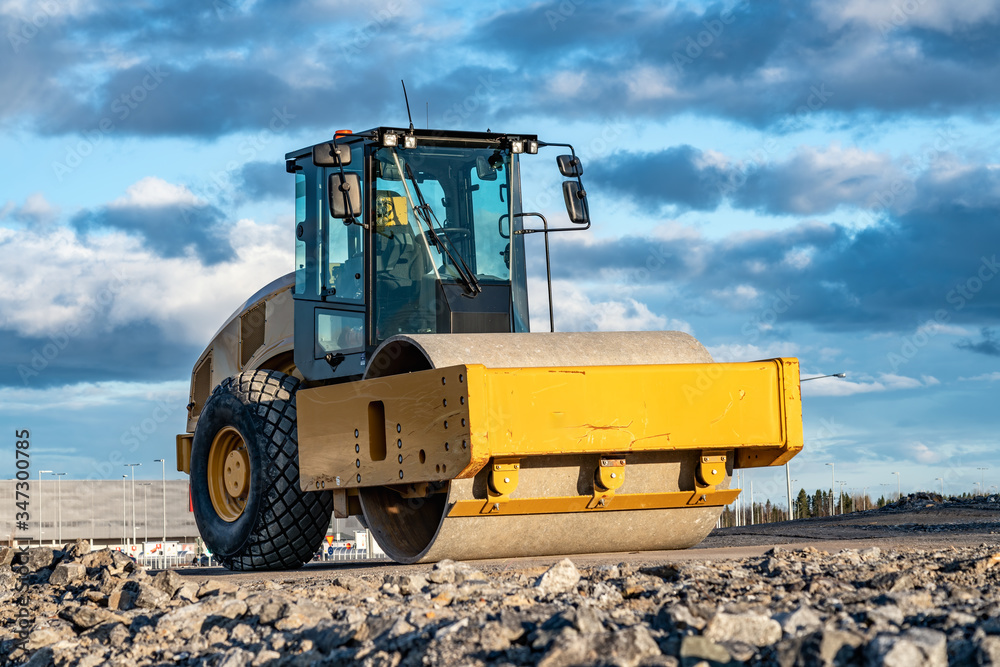 Eye catching yellow road roller with enclosed climate controlled cabin stands on not ready new road, stones, blue sky, clouds, front left side view. Clean shiny old heavy tractor Eye catching yellow road roller with enclosed climate controlled cabin stands on not ready new road, stones, blue sky, clouds, front left side view. Clean shiny old heavy tractor