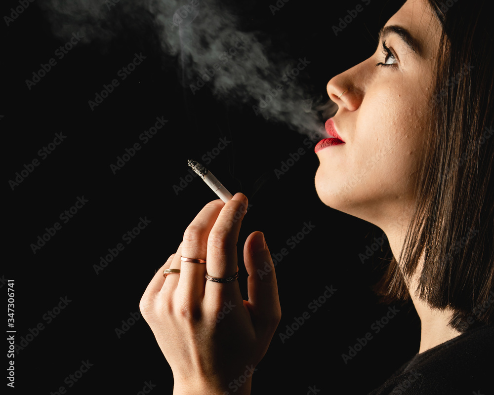Fototapeta premium Low key close-up young woman smoking cigarette on black background. Copy space for text message.