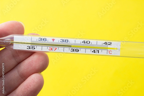 Mercury thermometer in hand on yellow background. Close up photo. Thermometer shows a temperature of 38.7 degrees Celsius photo